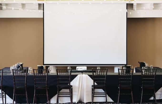 Wine And Culinary Events 4 A large screen is displayed in a room furnished with chairs and tables, ready for a presentation or meeting.